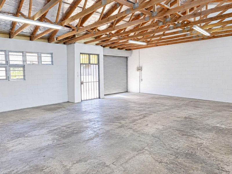 Spacious industrial garage featuring exposed timber roof trusses, concrete flooring, secure roller shutter door, and ample natural light through steel frame windows.