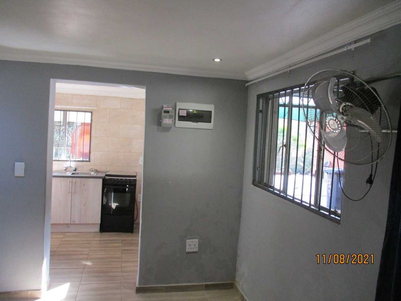 An open-plan living area featuring tiled floors, security bars on windows, and a visible electrical distribution board, leading into a functional kitchen space.
