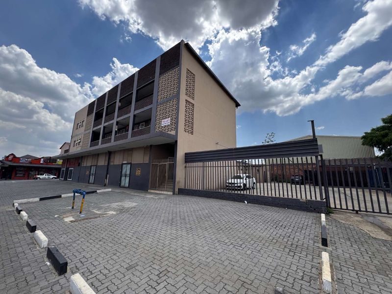 Exterior view of a multi-story mixed-use building featuring a paved parking area, secure metal fencing, and decorative breeze block architectural details.