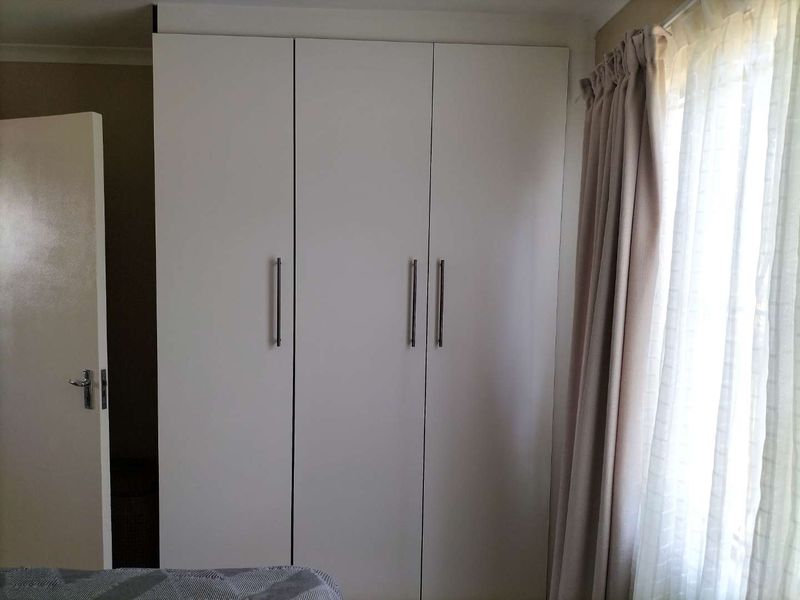 A bedroom featuring a clean, white built-in wardrobe with modern handles, positioned next to a window with curtains.