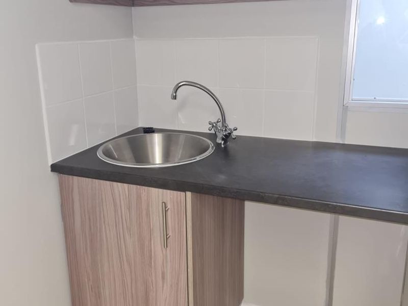 A compact kitchenette area featuring a stainless steel sink, laminate countertop, base cabinetry, and a wall-mounted shelf with tiled splashback.