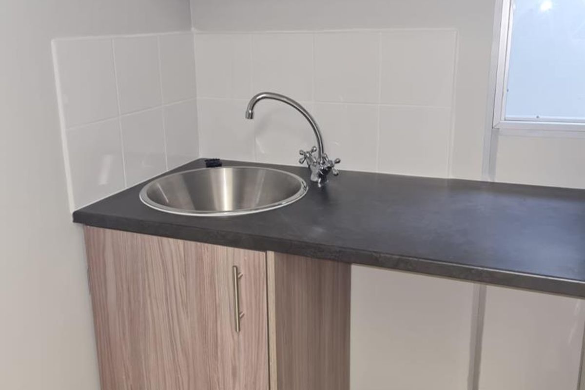 A compact kitchenette area featuring a stainless steel sink, laminate countertop, base cabinetry, and a wall-mounted shelf with tiled splashback.