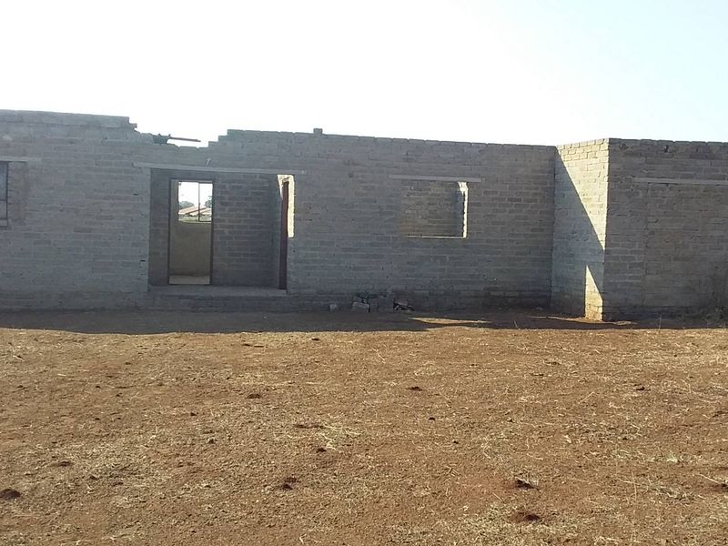An unfinished residential structure showing exposed brick walls and open window and door frames on a vacant plot of land.