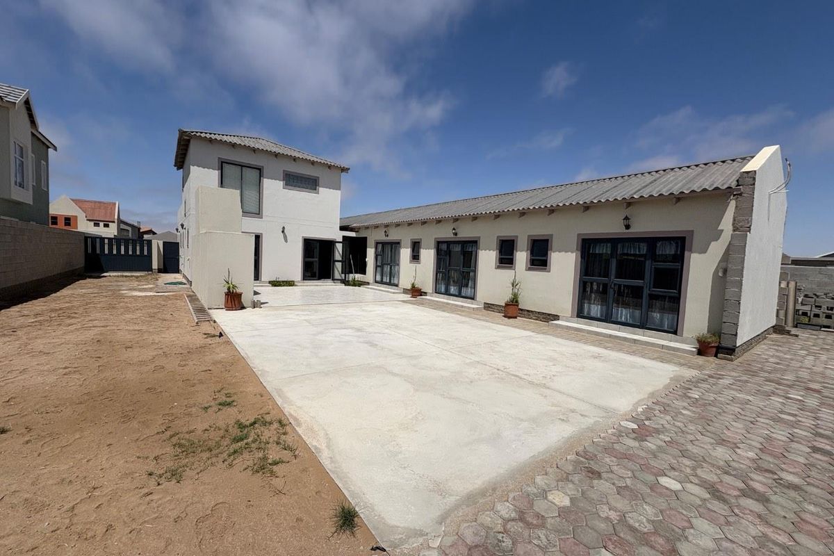 Rear view of the property featuring a large concrete patio area, paved driveway section, and multi-story building structure with dark-framed glass doors.