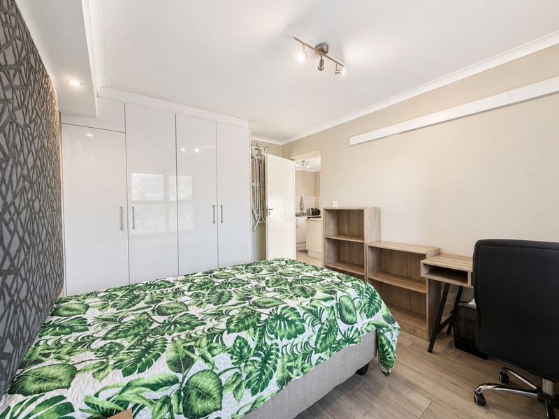A well-maintained bedroom featuring built-in white cabinetry, laminate flooring, and a textured feature wall, with direct access to the kitchen area.