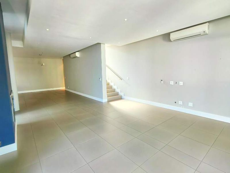 Spacious open-plan living area featuring neutral tiled flooring, recessed ceiling lighting, and a wall-mounted air conditioning unit, leading to an internal staircase.