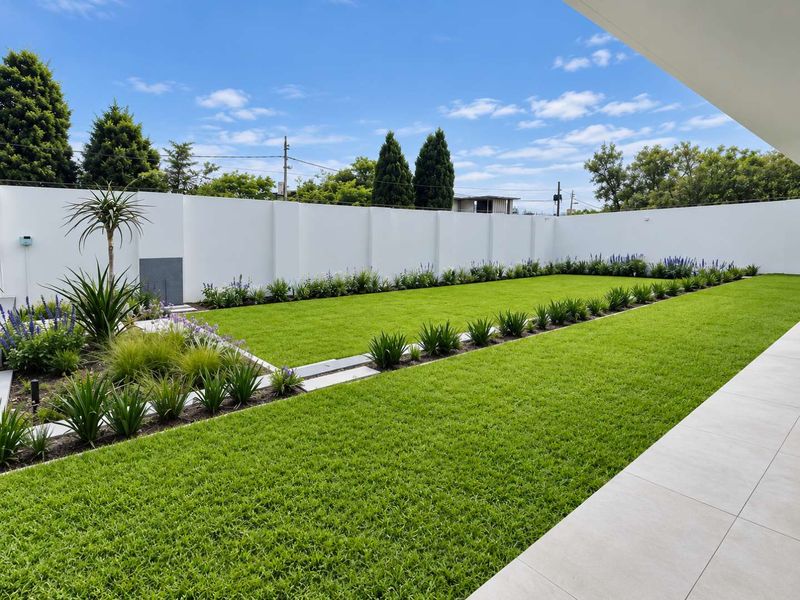 A well-maintained, landscaped garden featuring a manicured lawn, structured garden beds, and a clean perimeter boundary wall, viewed from a tiled patio area.