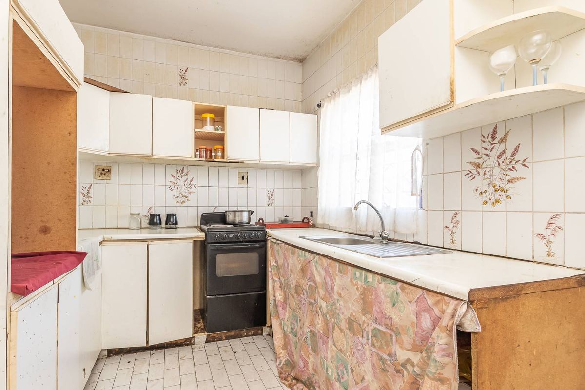 A dated kitchen featuring original tiled walls, built-in cabinetry, a stainless steel sink, and a freestanding stove, requiring significant renovation and modernization.