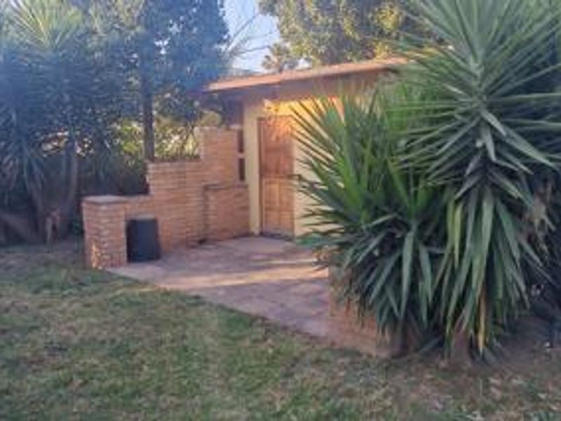 A rear exterior view featuring a paved patio area, brick wall structure, and a wooden door, surrounded by established garden vegetation.