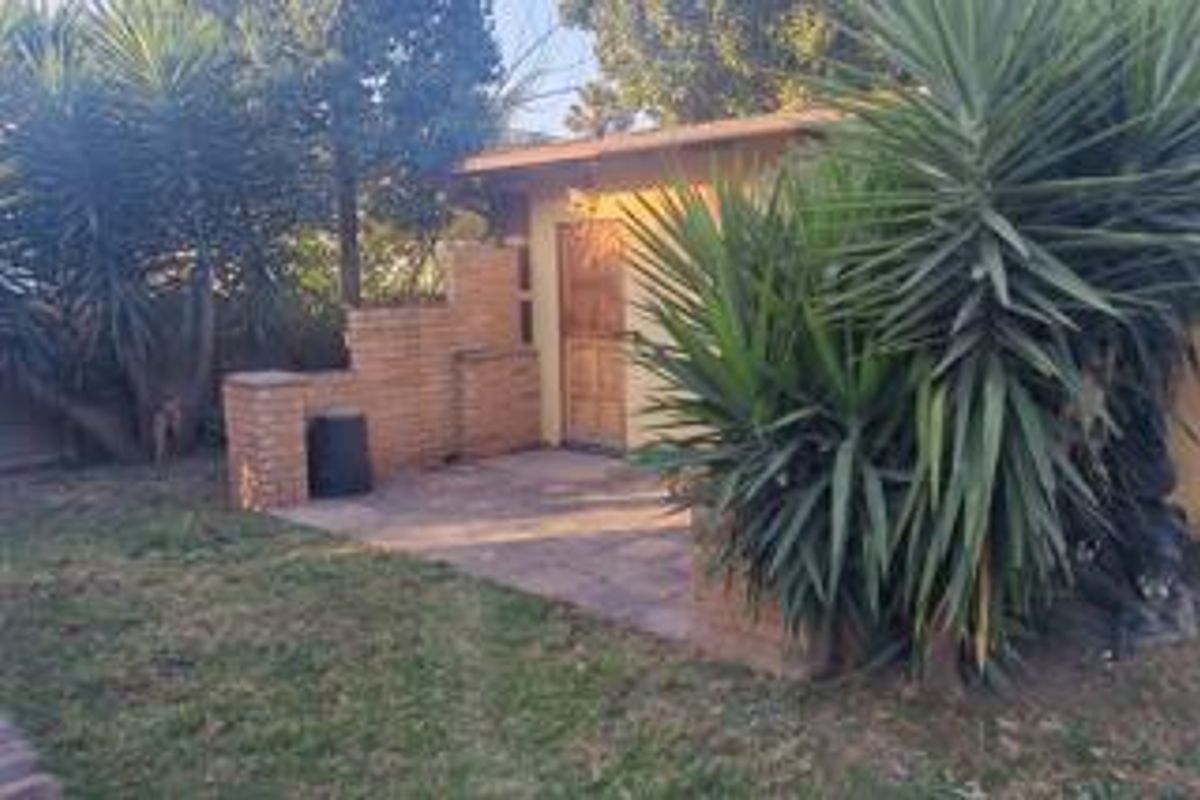 A rear exterior view featuring a paved patio area, brick wall structure, and a wooden door, surrounded by established garden vegetation.