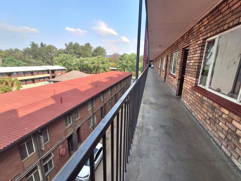 An exterior walkway balcony featuring exposed brick walls, concrete flooring, and a metal safety balustrade overlooking the apartment complex grounds.