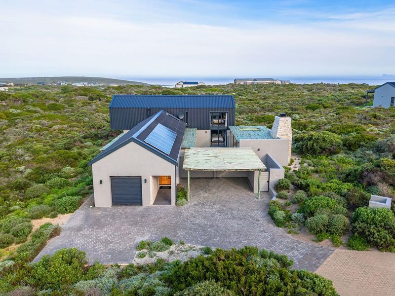 Aerial view of a modern coastal home featuring a metal roof, solar panels, stone chimney, and a paved driveway with a covered carport.