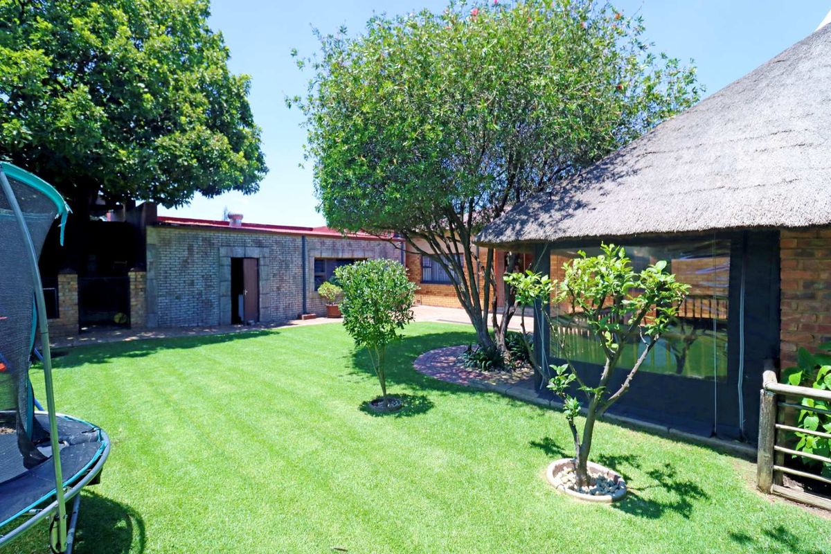 A well-maintained rear garden featuring a lush lawn, mature trees, and a thatched-roof outdoor structure with clear protective enclosures.