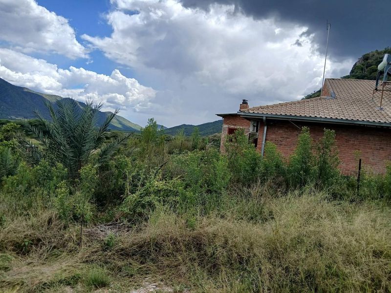 Rear view of a brick farmhouse set against a mountainous landscape, featuring a tiled roof and chimney, surrounded by overgrown vegetation.