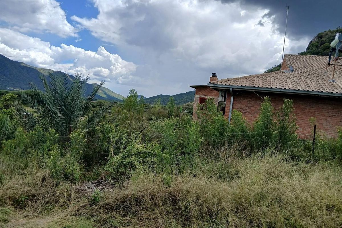 Rear view of a brick farmhouse set against a mountainous landscape, featuring a tiled roof and chimney, surrounded by overgrown vegetation.