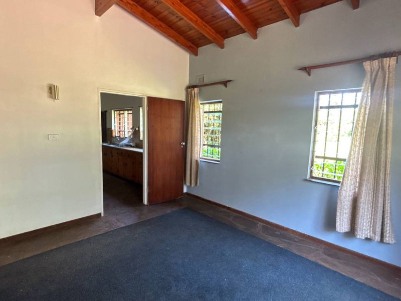 Spacious living area featuring exposed timber ceiling beams, large windows with security bars, and an arched entryway leading to the kitchen.