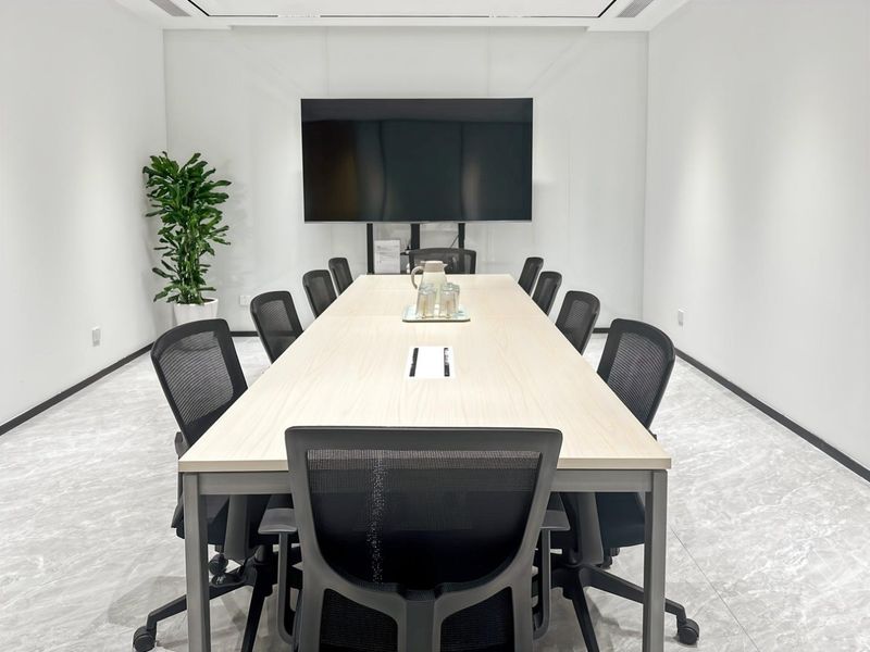 A modern, well-lit office boardroom featuring a large central meeting table, ergonomic seating, and a wall-mounted display screen.