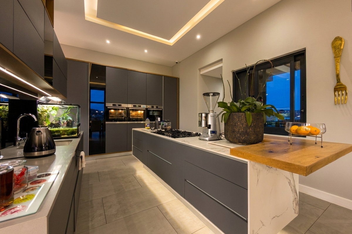 Modern kitchen featuring dark cabinetry, integrated ovens, a stone-topped island with a wooden breakfast bar, and recessed ceiling lighting.