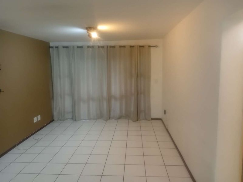 An empty living room featuring neutral tiled flooring, a single accent wall, and a large window covered by curtains.