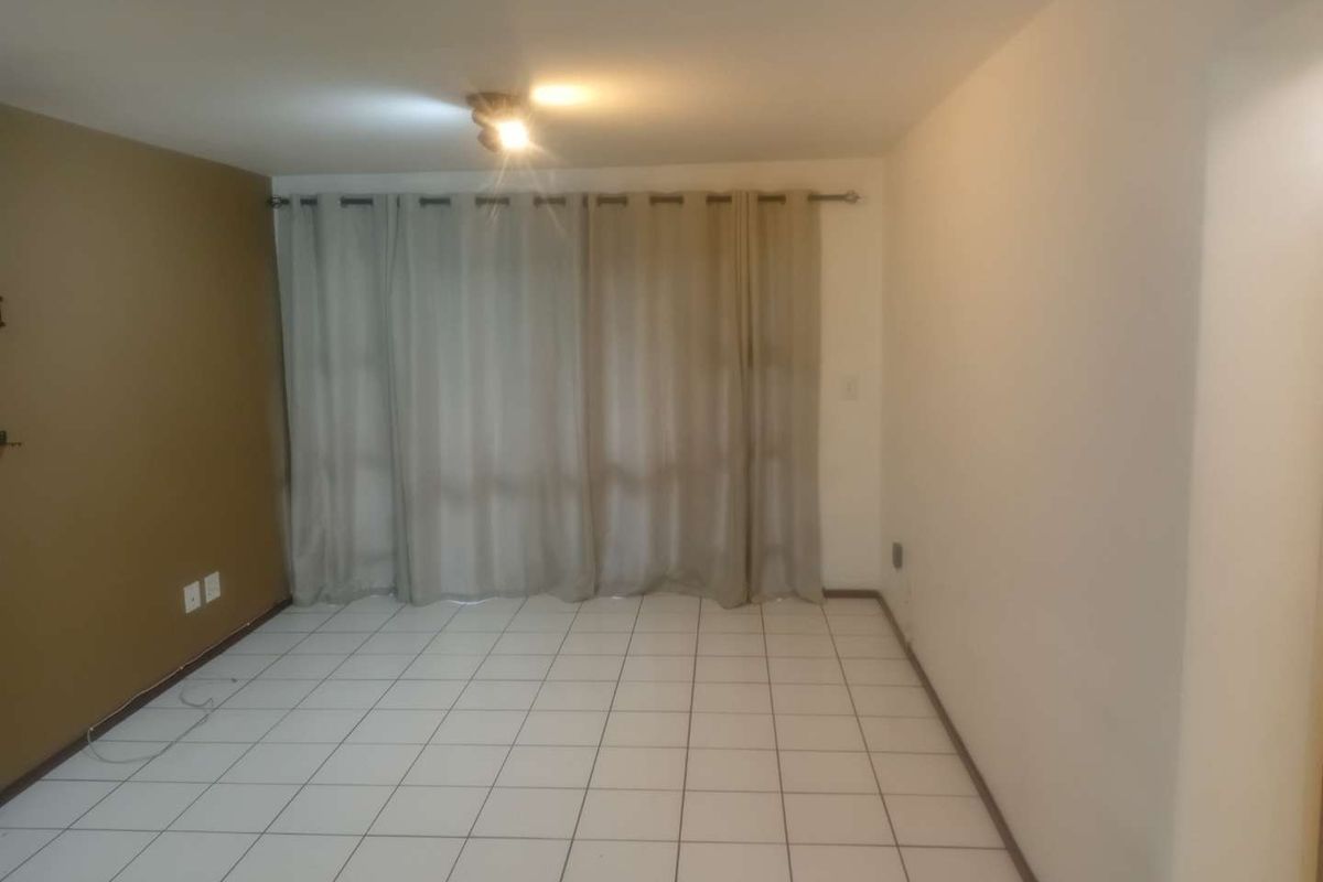 An empty living room featuring neutral tiled flooring, a single accent wall, and a large window covered by curtains.