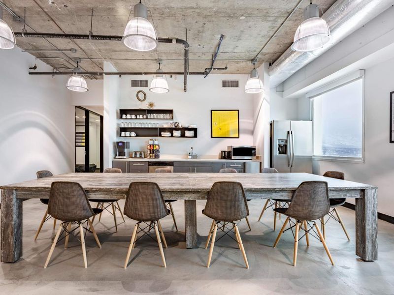 Modern open-plan office space featuring an industrial aesthetic with exposed concrete ceilings, polished concrete floors, and a functional kitchenette area.