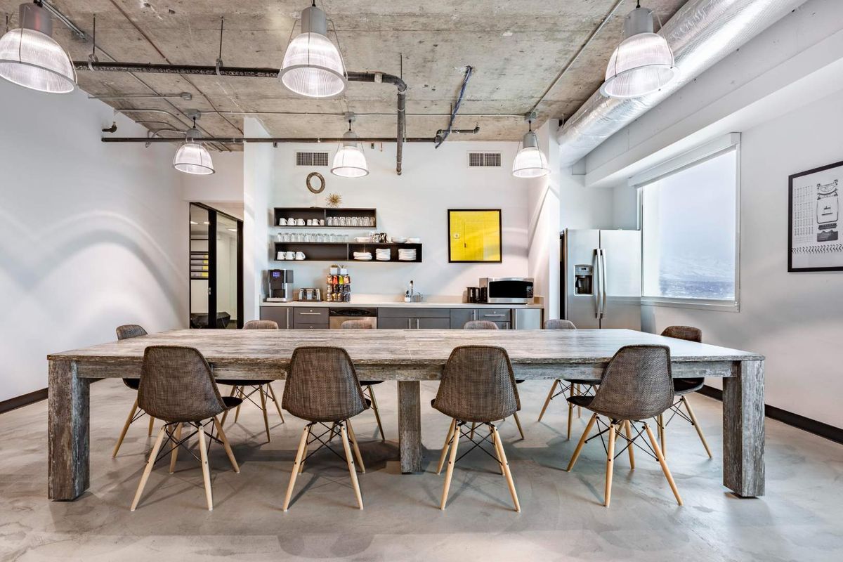 Modern open-plan office space featuring an industrial aesthetic with exposed concrete ceilings, polished concrete floors, and a functional kitchenette area.