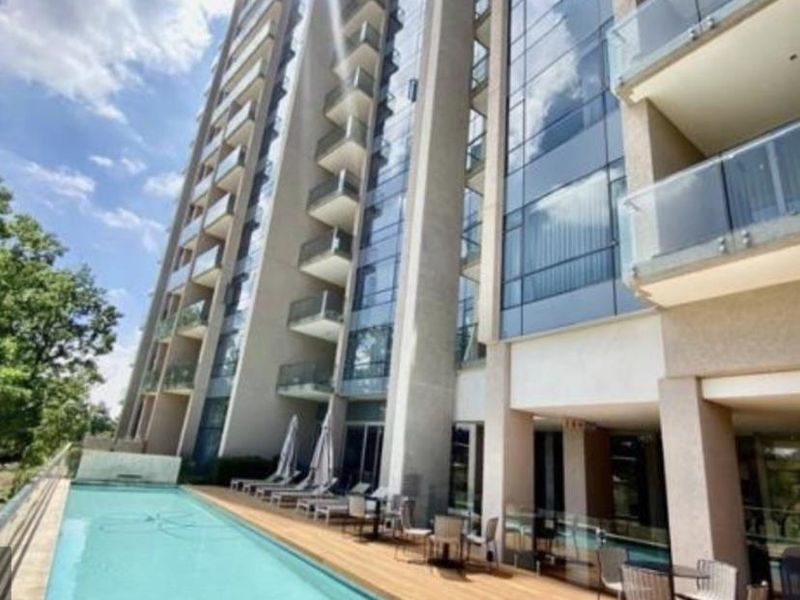 Modern apartment complex featuring a swimming pool with timber decking and glass balustrades, set against a contemporary glass-facade building.