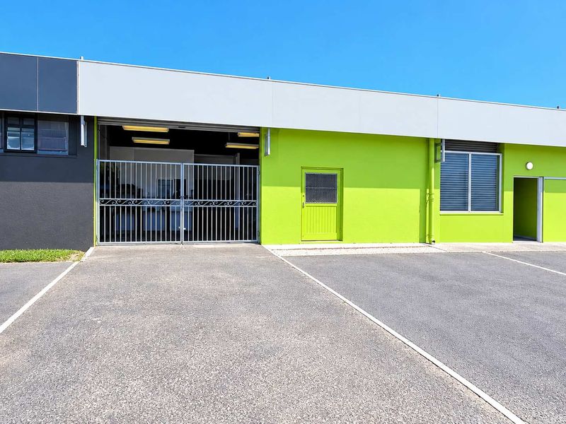 Front elevation of an industrial unit featuring a roller shutter door, security gate, and asphalt parking area.