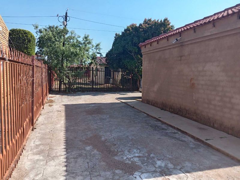 A rear exterior area featuring concrete paving, metal perimeter fencing, and a tiled roof structure with visible wear on the wall surfaces.