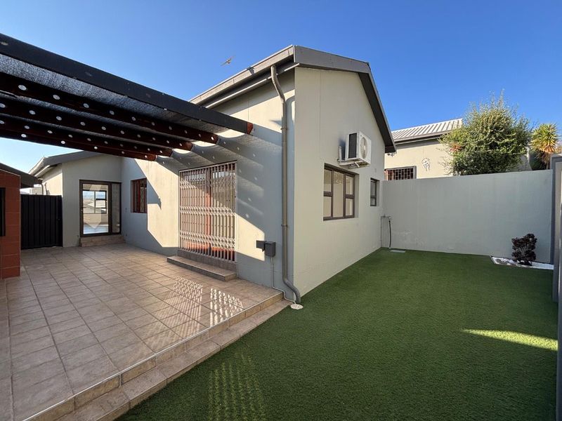 Rear exterior view of the townhouse featuring a tiled patio with a pergola, artificial lawn, and secure boundary walling.