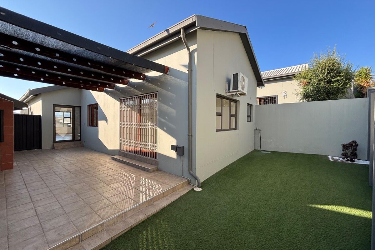 Rear exterior view of the townhouse featuring a tiled patio with a pergola, artificial lawn, and secure boundary walling.