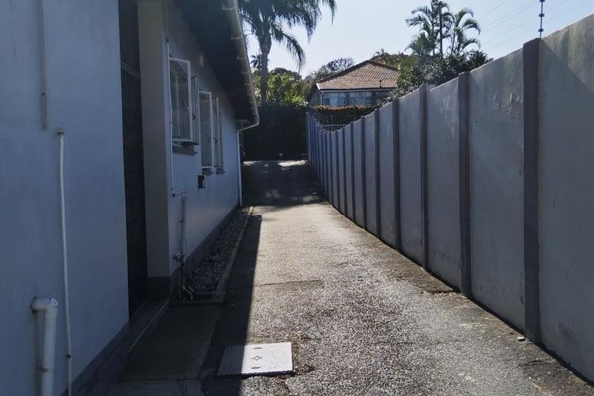 A narrow rear exterior pathway featuring an asphalt driveway, concrete boundary wall, and drainage infrastructure along the side of the apartment building.