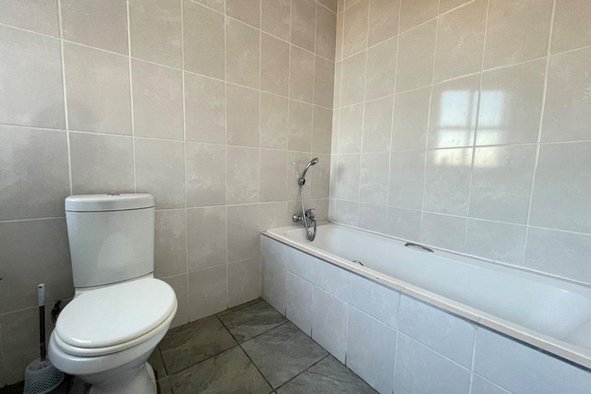 A functional bathroom featuring a standard bathtub with a handheld shower mixer, a toilet, and neutral wall and floor tiling.
