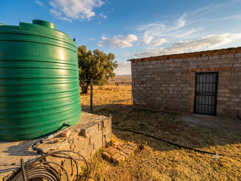 Exterior view of a farm utility area featuring a large green water storage tank on a concrete base next to a cinder block outbuilding.