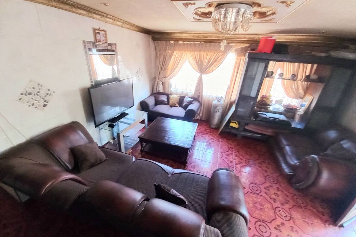 A living room featuring patterned floor tiles, decorative ceiling molding with a central chandelier, and large windows allowing natural light.