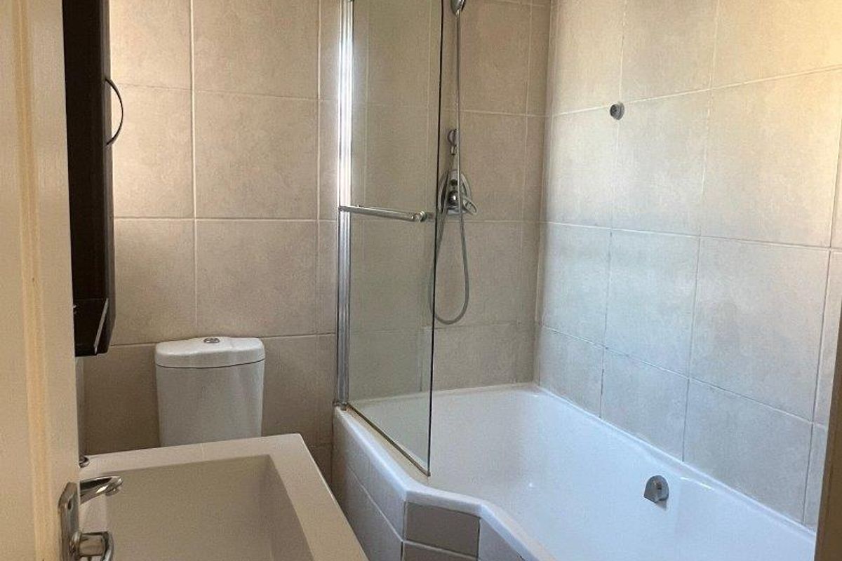 A clean, functional bathroom featuring a built-in vanity, toilet, and a bathtub with an integrated glass shower screen and neutral wall tiling.