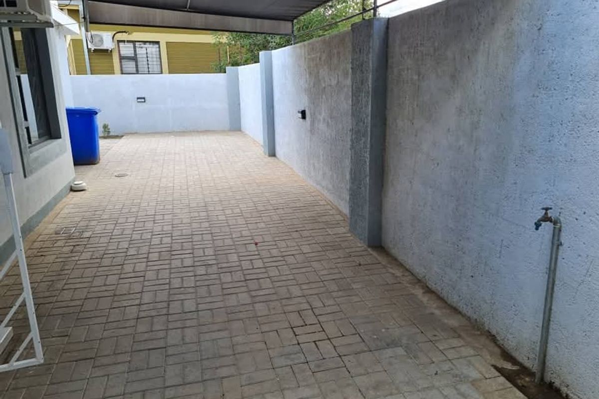 A paved side yard or exterior utility area featuring a covered patio section, boundary wall, and an external water tap.