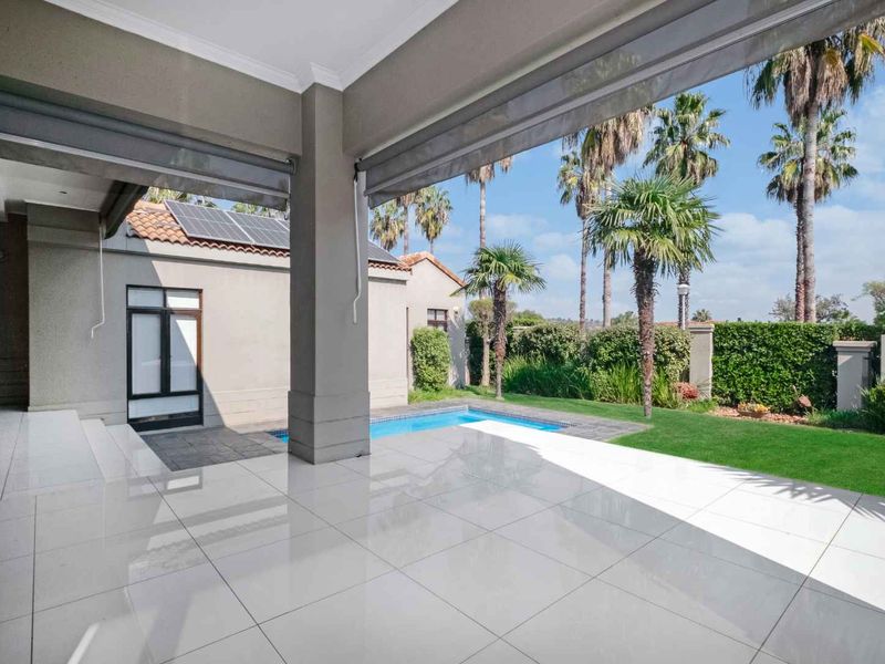 A spacious covered patio with tiled flooring, overlooking a private swimming pool and well-maintained garden area with mature palm trees.