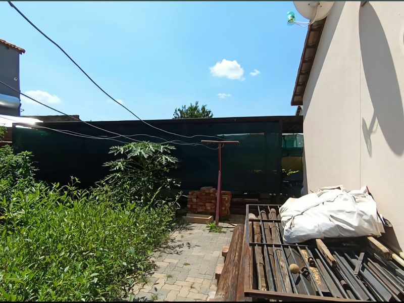 Rear exterior area featuring a paved walkway, shade cloth perimeter fencing, and established garden vegetation, requiring maintenance and clearing.