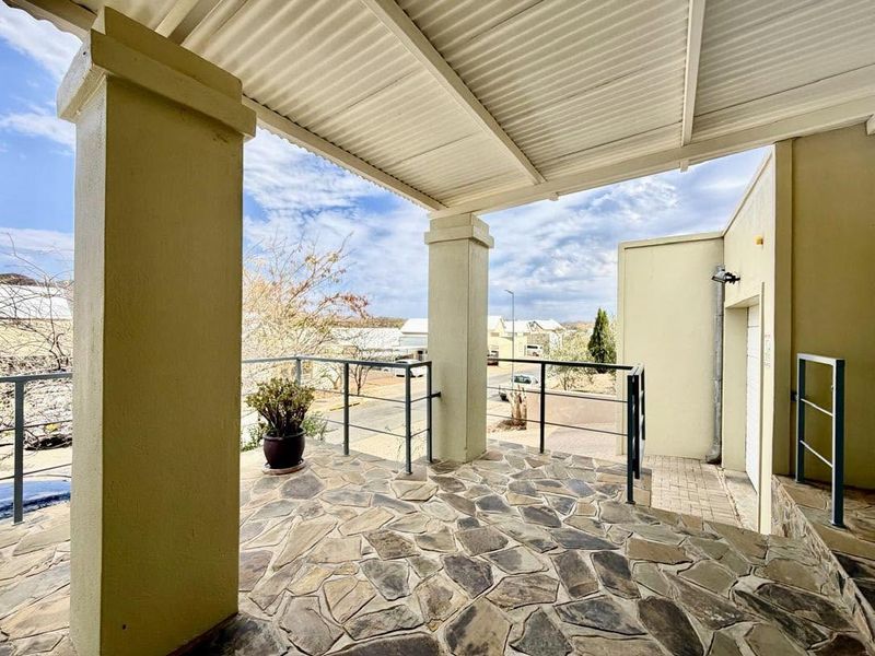A covered outdoor patio featuring stone flooring, concrete support pillars, and metal railings, offering an open view of the surrounding neighborhood.
