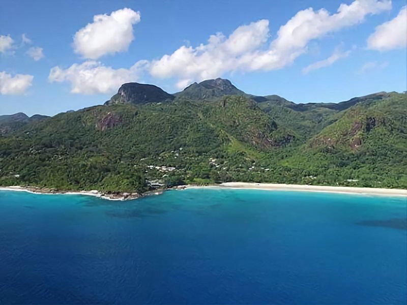 An aerial view of the coastal landscape featuring lush green mountains, a sandy beach, and clear blue ocean waters.