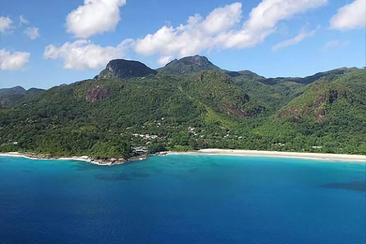 An aerial view of the coastal landscape featuring lush green mountains, a sandy beach, and clear blue ocean waters.