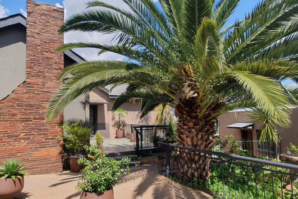 A well-maintained exterior rear area featuring a stone-clad chimney, paved patio space, and mature palm tree with metal balustrade fencing.