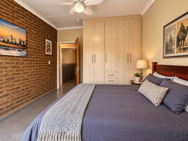 Spacious bedroom featuring a warm exposed brick feature wall, ample built-in cupboard storage, tiled flooring, and a ceiling fan for comfort.