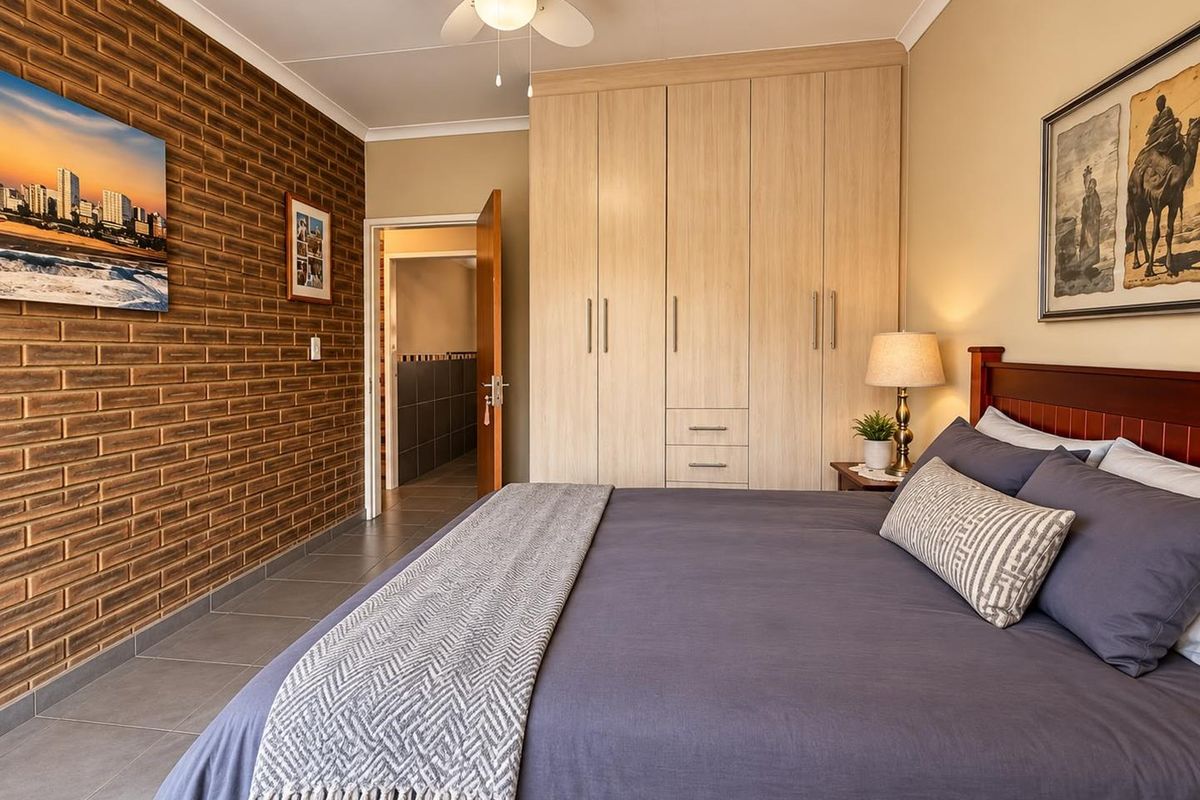 Spacious bedroom featuring a warm exposed brick feature wall, ample built-in cupboard storage, tiled flooring, and a ceiling fan for comfort.