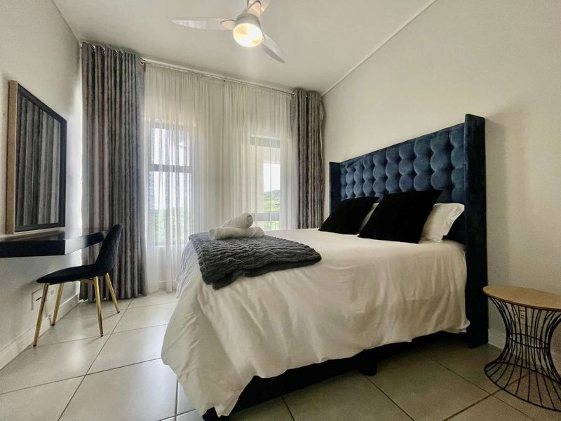 A well-maintained bedroom featuring tiled flooring, a ceiling fan, and large windows with floor-to-ceiling curtains, providing a bright and comfortable living space.