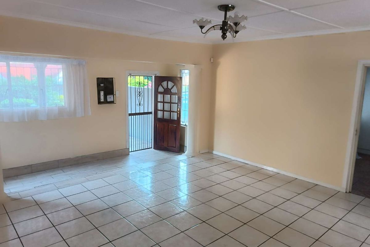 A spacious, empty living area featuring tiled flooring, a decorative ceiling light fixture, and a wooden entrance door with security gate.