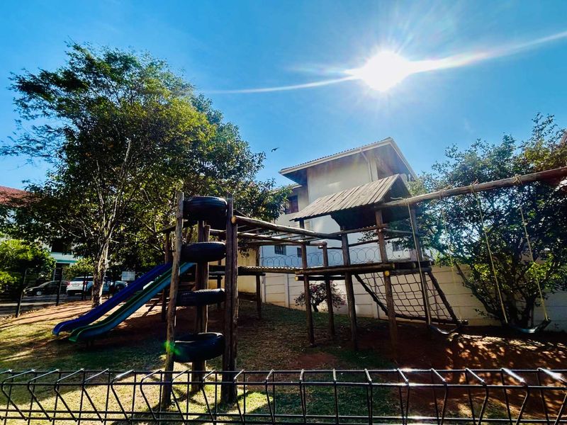Outdoor communal garden area featuring a wooden playground structure with slides and swings, enclosed by perimeter fencing.
