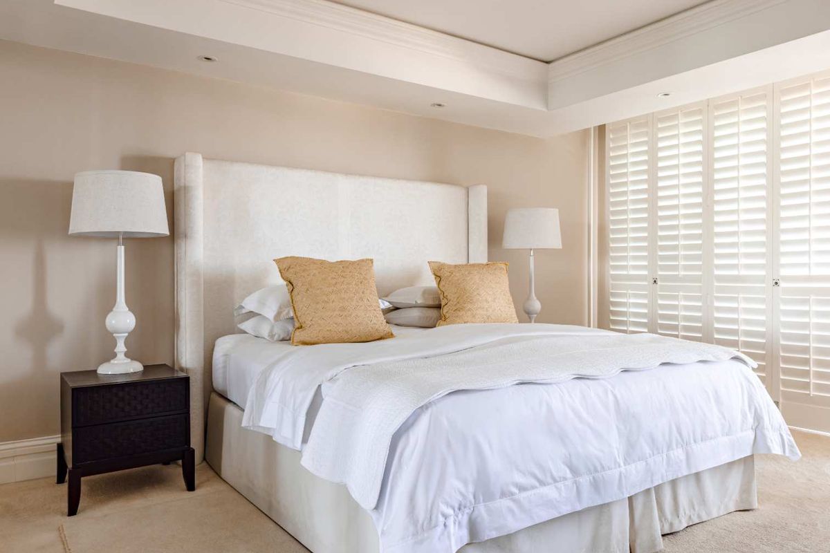 A well-appointed bedroom featuring a recessed ceiling with integrated downlights and classic plantation shutters, offering a clean and neutral aesthetic.