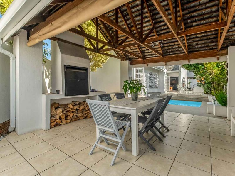 Covered outdoor patio featuring a built-in braai, exposed timber roof trusses, and tiled flooring, overlooking the swimming pool and garden area.